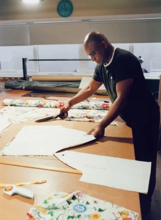 An adult male cuts fabric with scissors in a bright room. Floral pattern fabric is spread across a large table. Interior daytime.