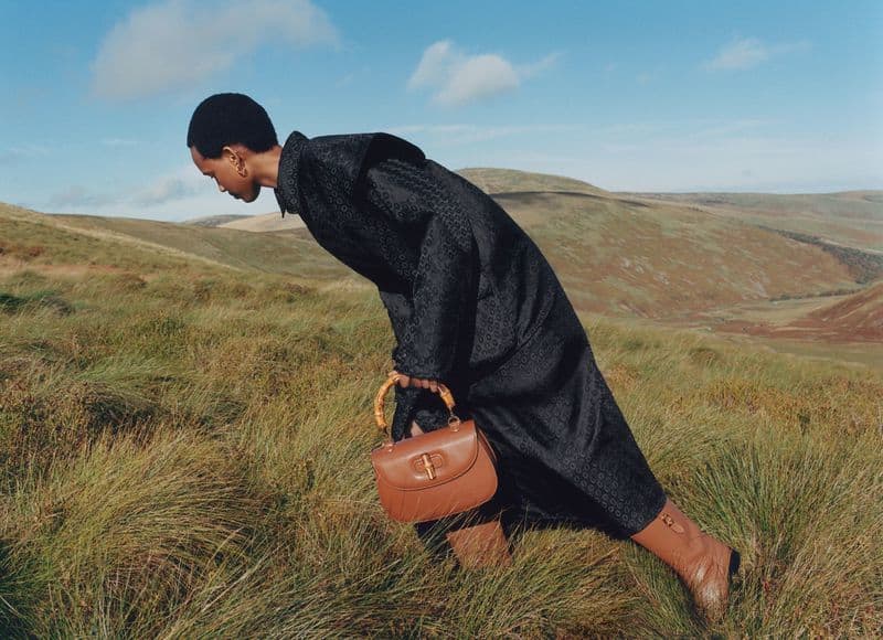 Woman in a patterned coat and tan boots with a brown handbag posing in a grassy field with rolling hills and blue sky.