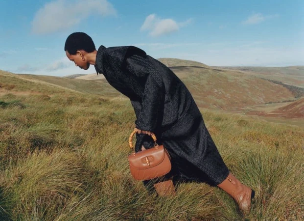 Woman in a patterned coat and tan boots with a brown handbag posing in a grassy field with rolling hills and blue sky.