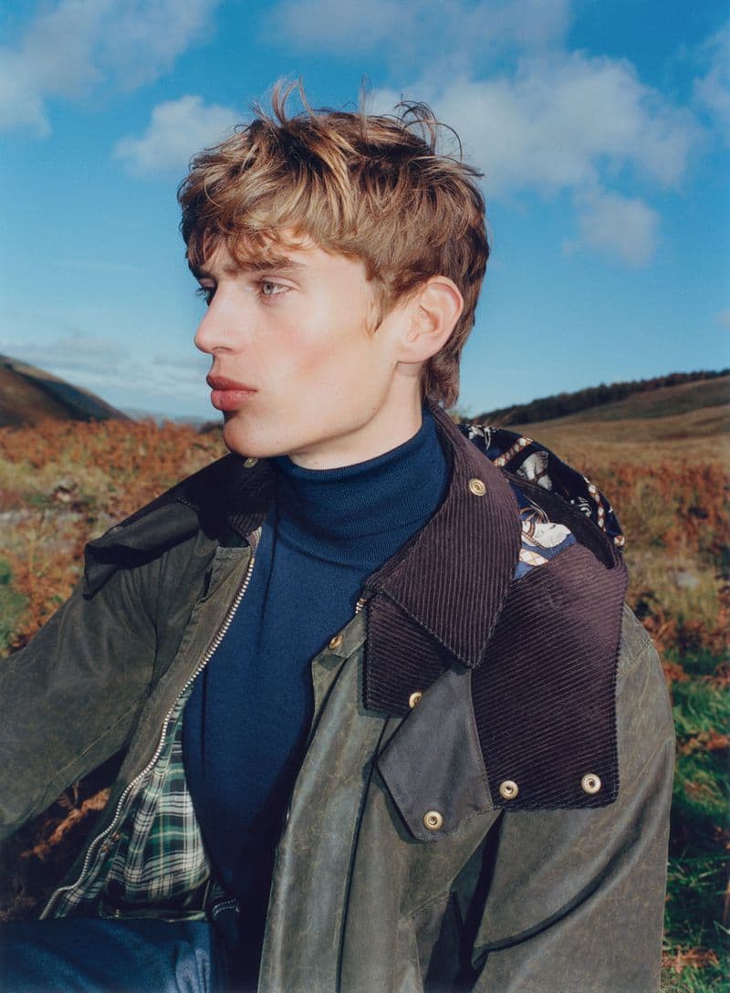 Male model with blonde hair and blue turtleneck sweater in a brown jacket posing in outdoor rural setting on a cloudy day.