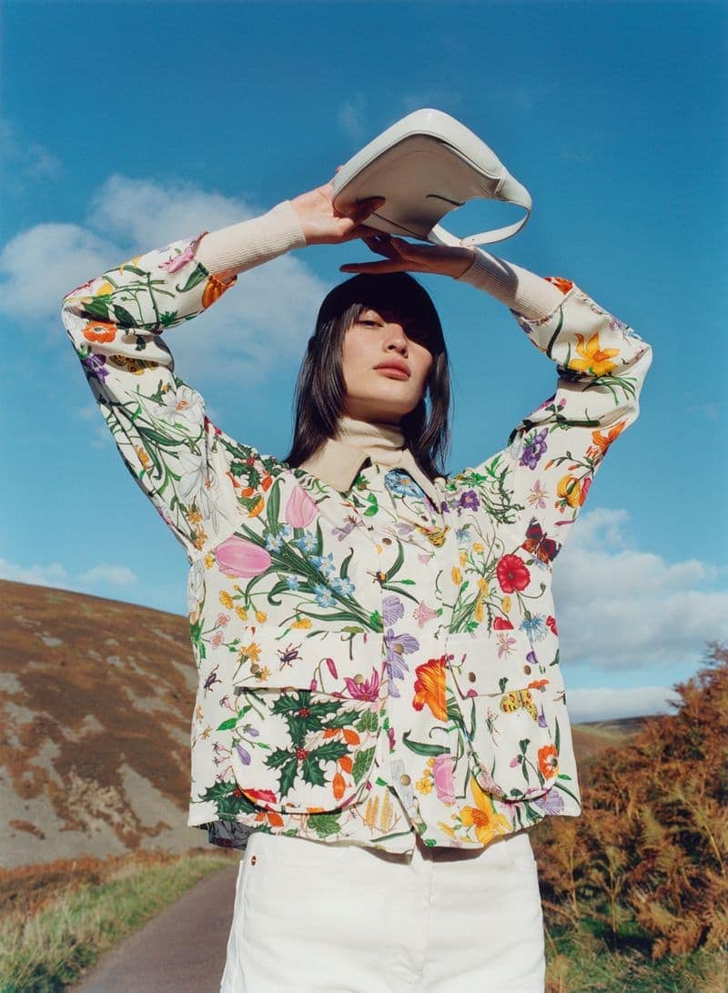 Young woman in floral jacket holding a handbag outdoors against a blue sky and rural background, creating a fashion editorial image.