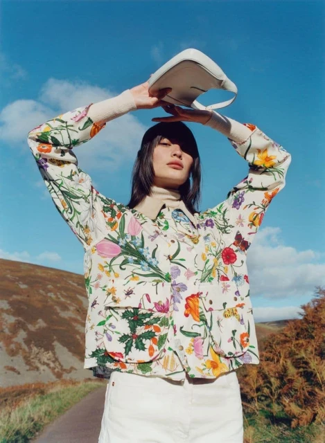 Young woman in floral jacket holding a handbag outdoors against a blue sky and rural background, creating a fashion editorial image.