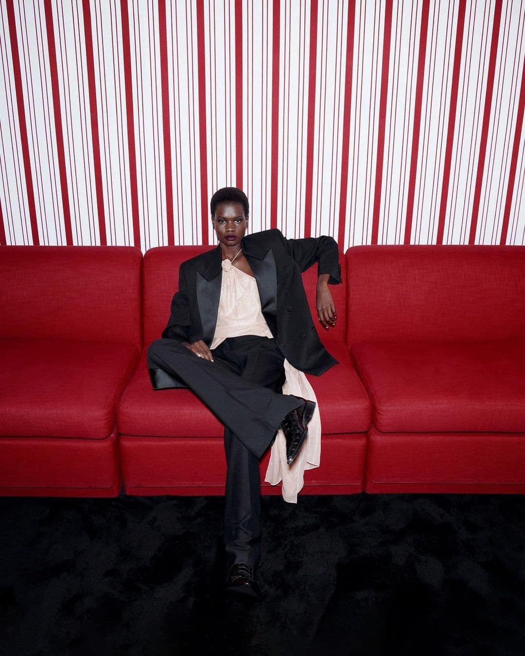A black female model with short hair sits elegantly on a red couch wearing a black tuxedo, satin top, and patent-leather shoes against a striped wall.