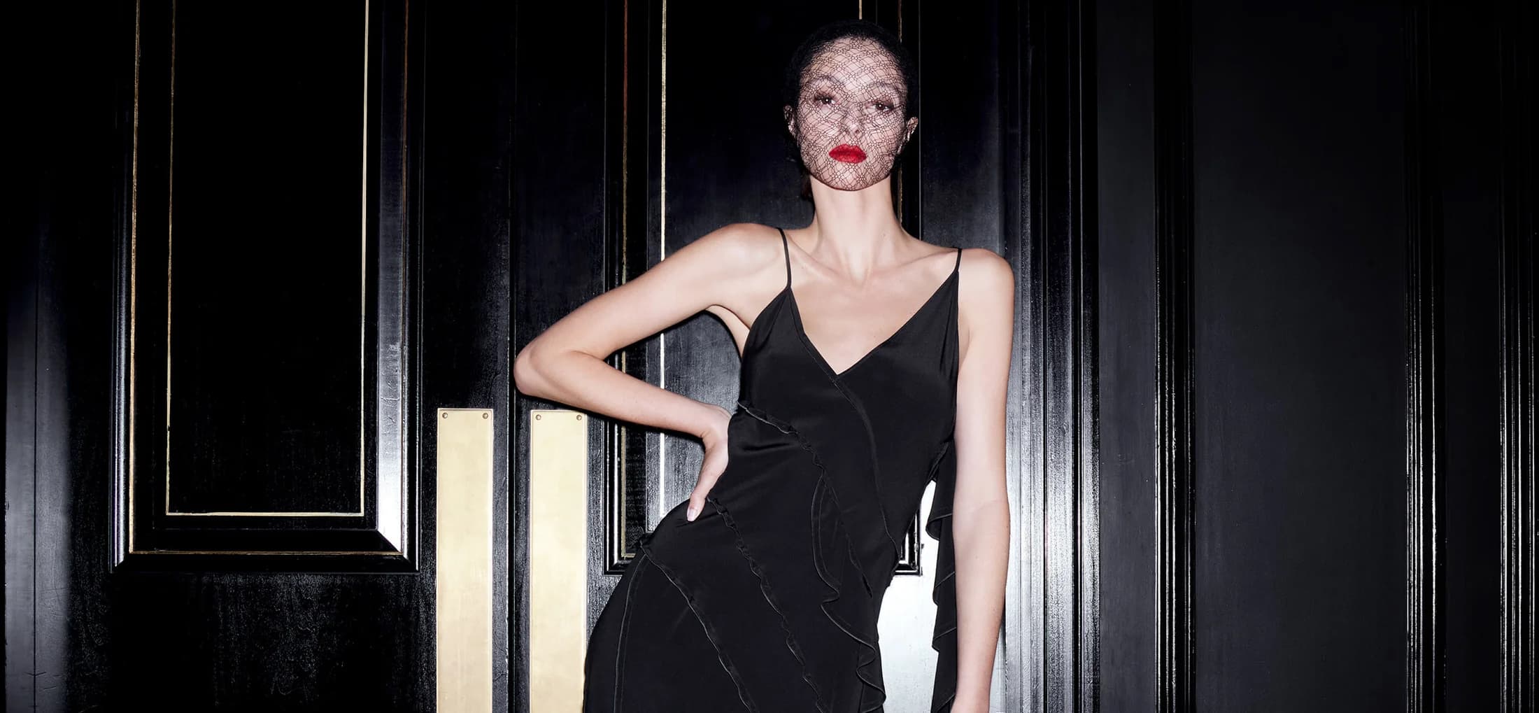 A woman in a black dress poses against a black door with gold trim, wearing a net veil and red lipstick, under studio lighting.