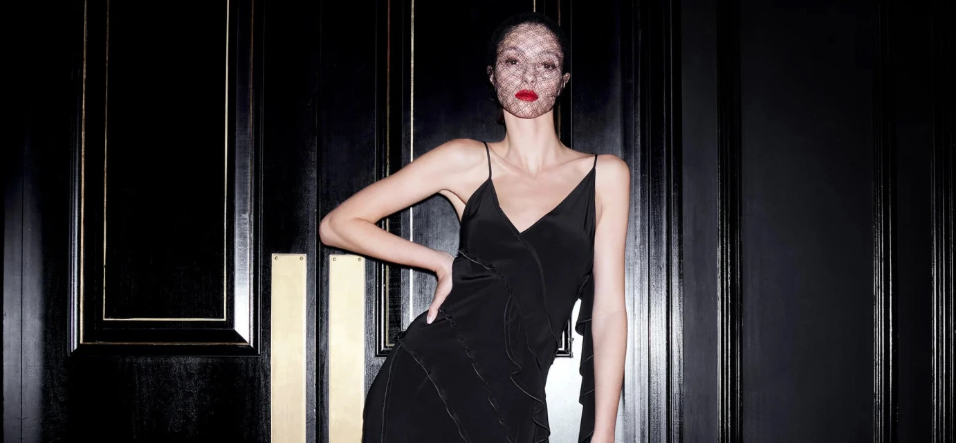 A woman in a black dress poses against a black door with gold trim, wearing a net veil and red lipstick, under studio lighting.