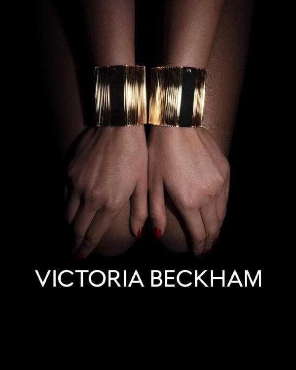 Woman's wrists adorned with gold cuff bracelets, juxtaposed against a stark black backdrop, with the words VICTORIA BECKHAM displayed.
