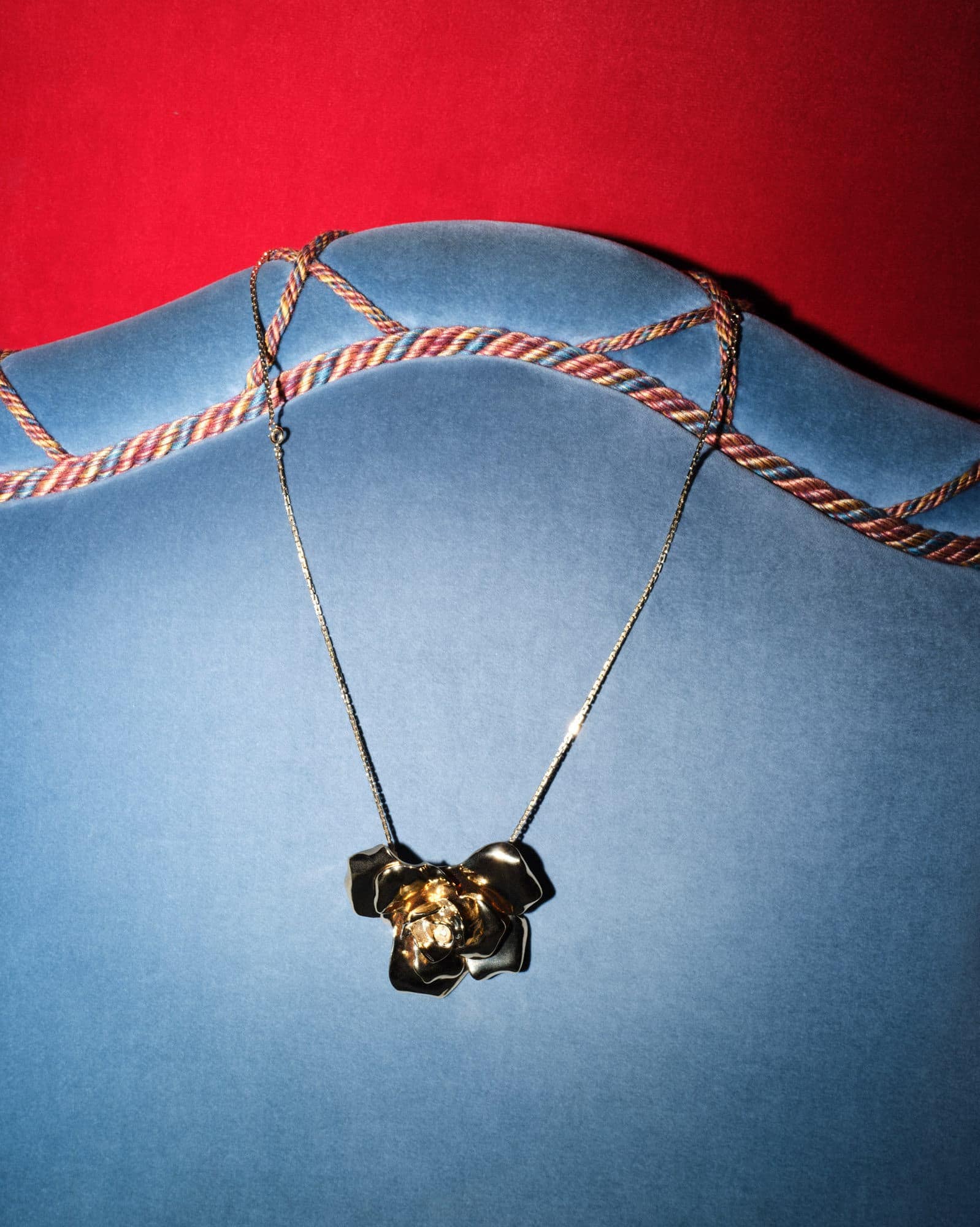 Necklace with gold-toned flower pendant on a delicate silver chain, draped on a light blue velvet chair against a red background.