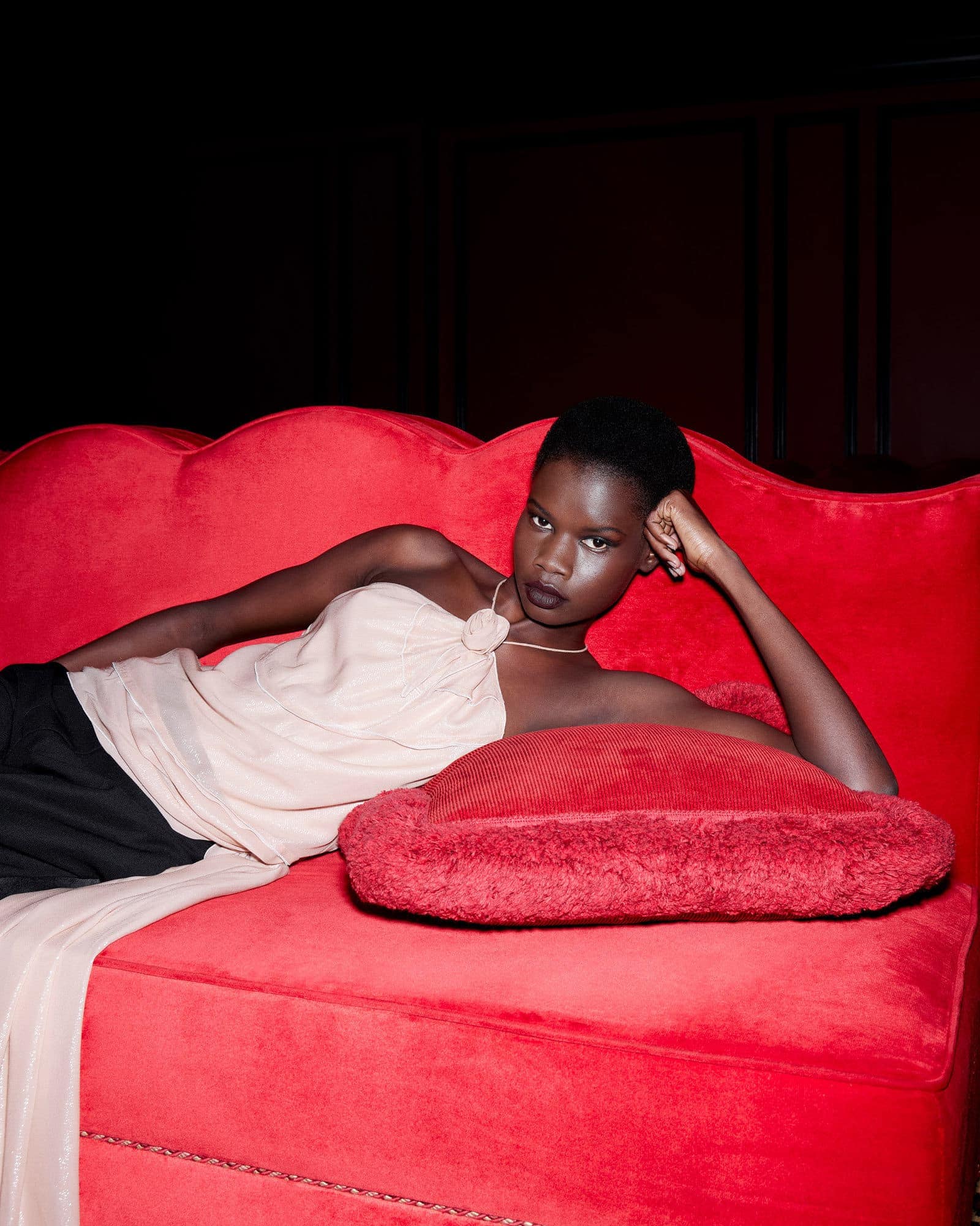 Elegant dark-skinned woman lounging on a bright red velvet couch wearing a beige top and black pants in a dark, moody indoor setting.