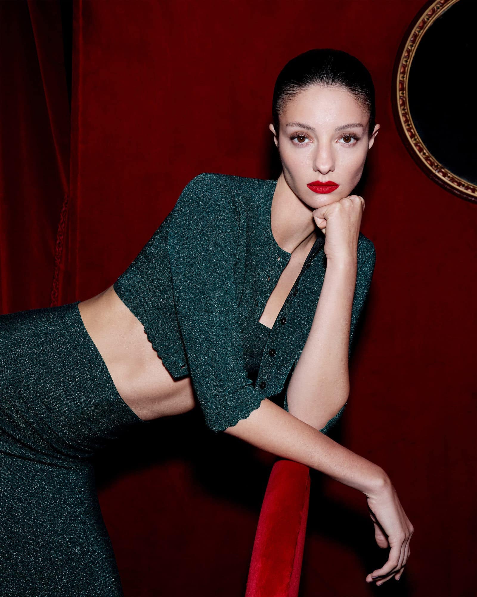 Model in a dark green sparkly cardigan leaning against red fabric with a round mirror on the wall