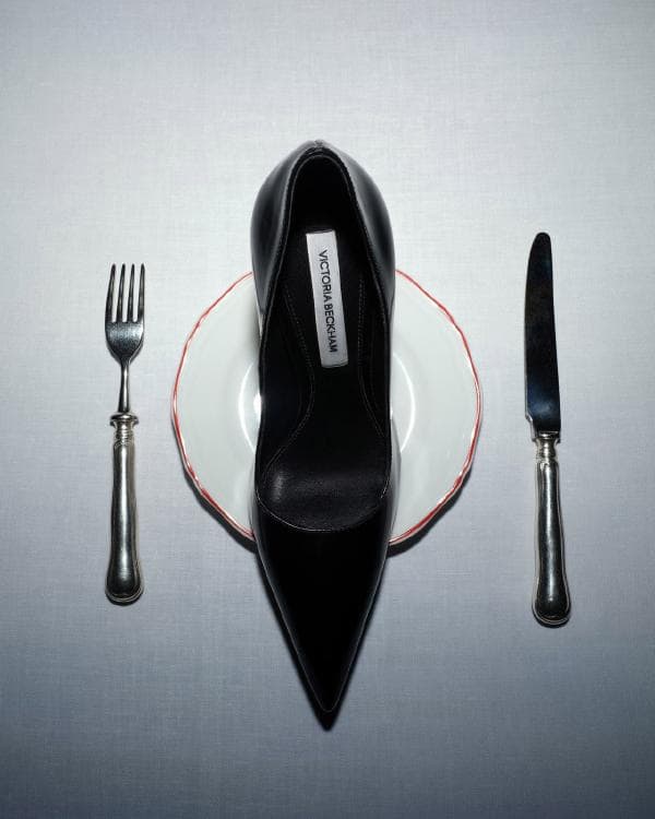 Overhead shot of a black stiletto heel shoe placed on a white plate with silverware, set against a white tablecloth backdrop.