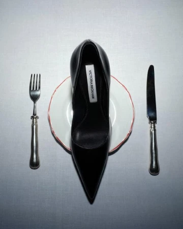 Overhead shot of a black stiletto heel shoe placed on a white plate with silverware, set against a white tablecloth backdrop.