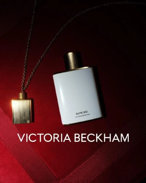 Victoria Beckham Suite 302 perfume bottle with gold pendant necklace against a dark red background with geometric fabric folds.