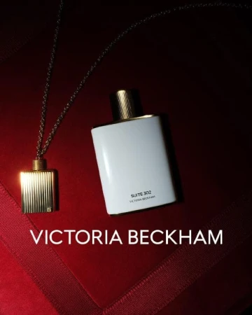 Victoria Beckham Suite 302 perfume bottle with gold pendant necklace against a dark red background with geometric fabric folds.