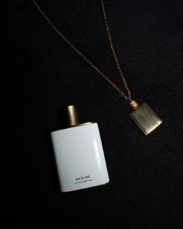 Flat lay studio shot of a white perfume bottle with a golden cap next to a gold necklace with a rectangular pendant.