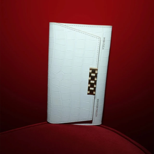 White textured clutch with gold hardware on a red fabric surface in a studio setting. Fashion accessory with visible logo.