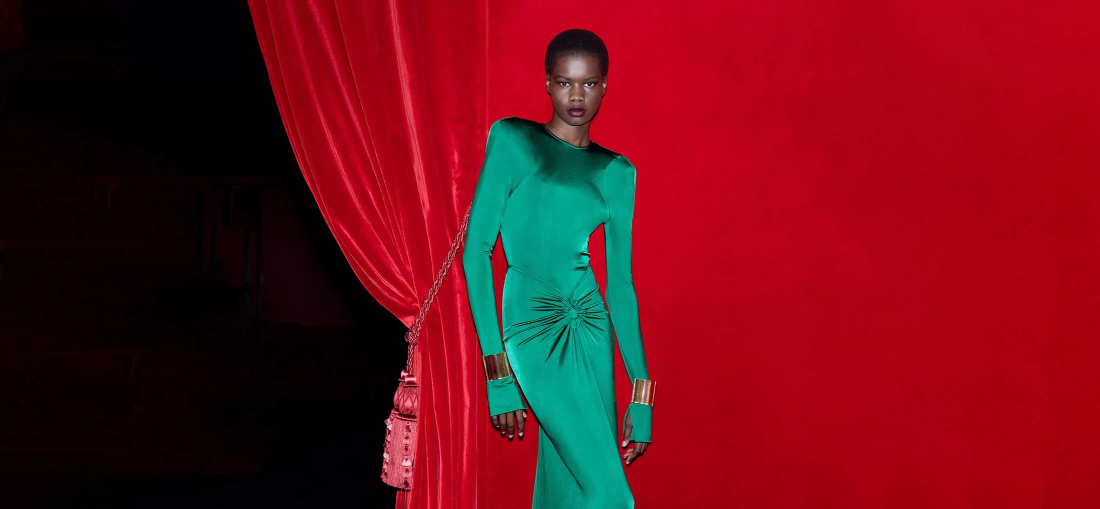 A Black woman with short hair poses in a long-sleeve green dress with gold cuffs in front of a red background with red velvet curtains.