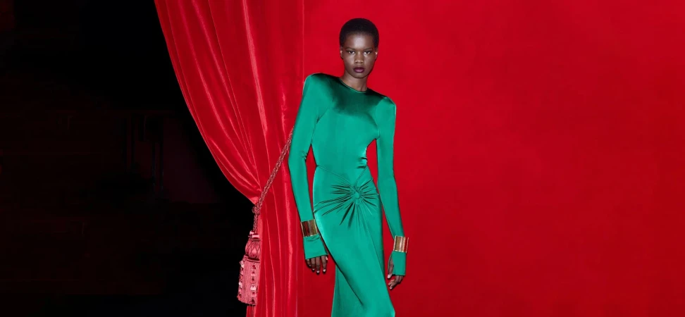 A Black woman with short hair poses in a long-sleeve green dress with gold cuffs in front of a red background with red velvet curtains.