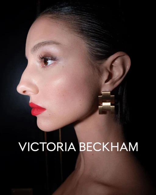 Profile portrait of fair-skinned adult female with red lipstick and gold geometric earrings, against a dark background.