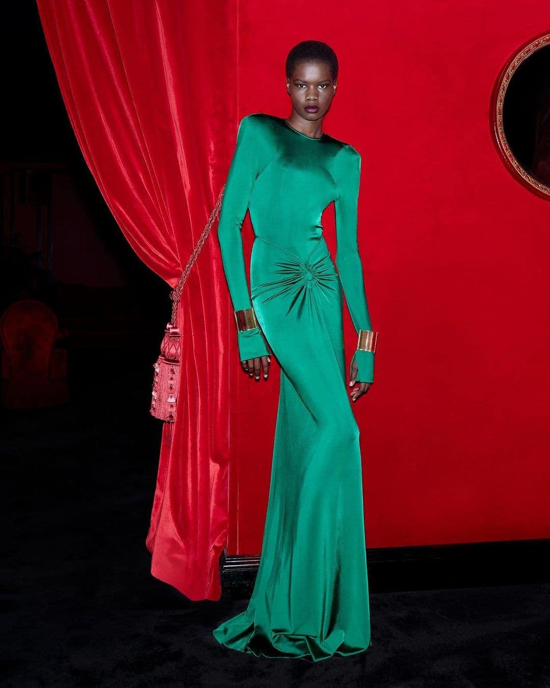 Elegant African woman in a long-sleeved green gown and gold cuffs poses against a red backdrop with a draped curtain and ornate frame.