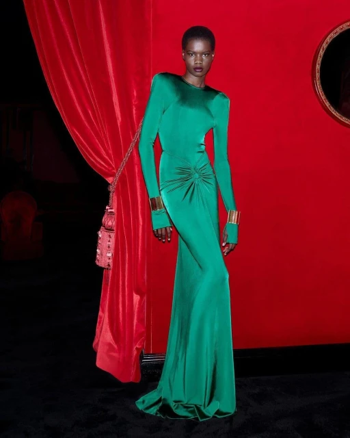 Elegant African woman in a long-sleeved green gown and gold cuffs poses against a red backdrop with a draped curtain and ornate frame.