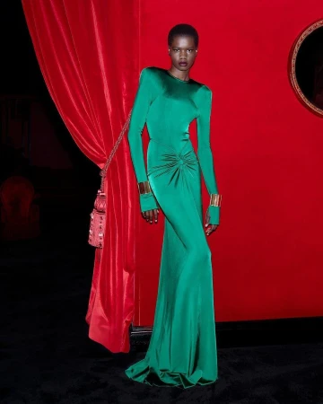 Elegant African woman in a long-sleeved green gown and gold cuffs poses against a red backdrop with a draped curtain and ornate frame.