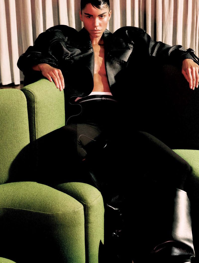 Fashionable adult woman in a black jacket and boots poses while sitting on a green sofa against a curtained backdrop indoors.
