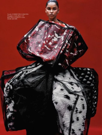 Woman in avant-garde fashion with oversized red, black, and white garment against a red backdrop, posing for a studio shot.