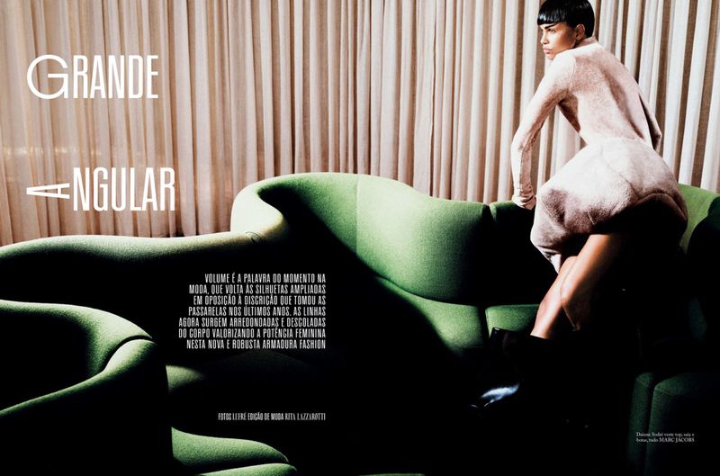 Fashion editorial shot of a woman in a light pink dress seated on a vintage green couch in front of neutral-toned curtain backdrop.