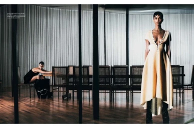 Model standing in a yellow dress in foreground with another model seated at a long table in the background behind glass panels