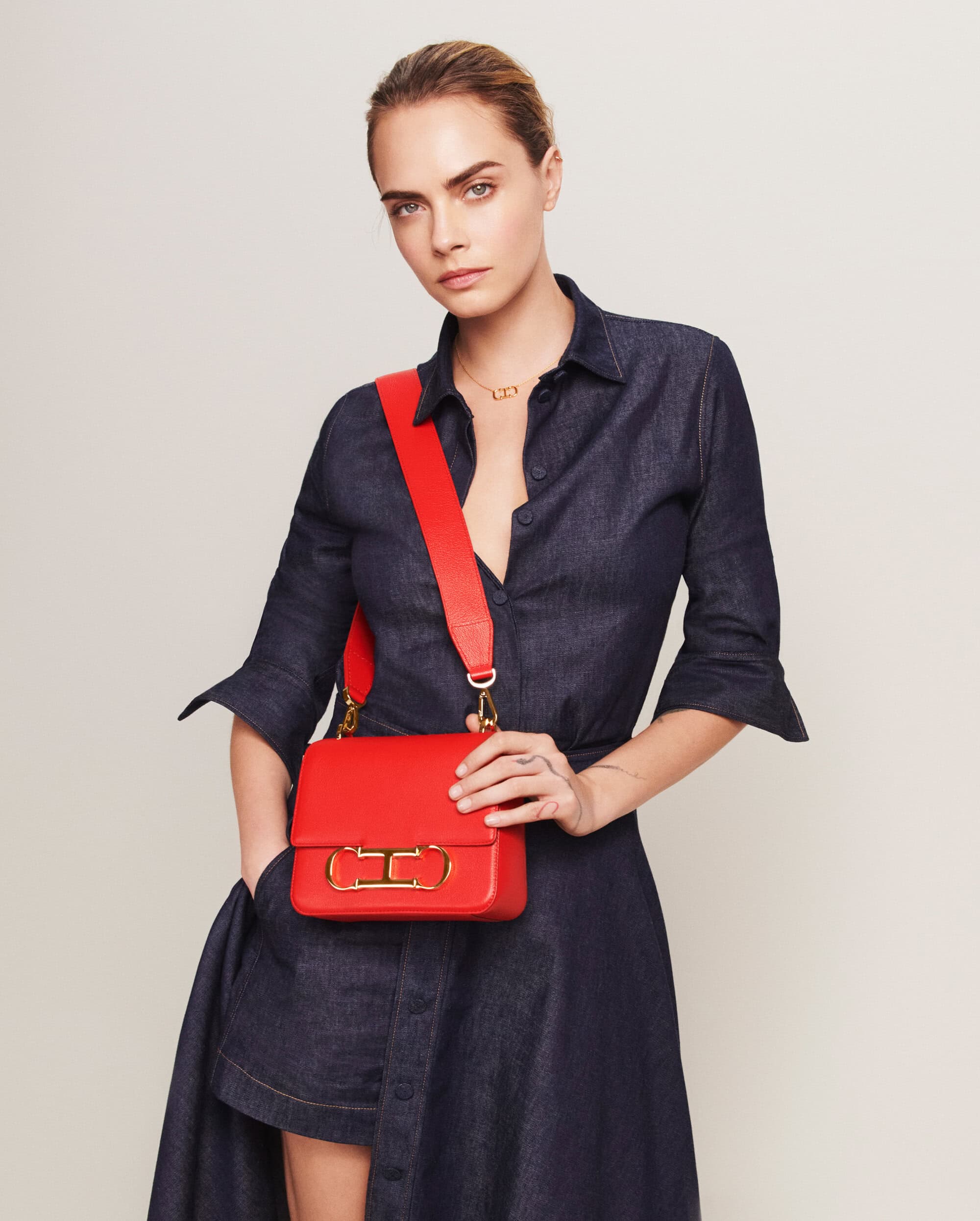 Fashion shot of woman in dark denim dress wearing red leather shoulder bag with gold hardware against an off-white background.