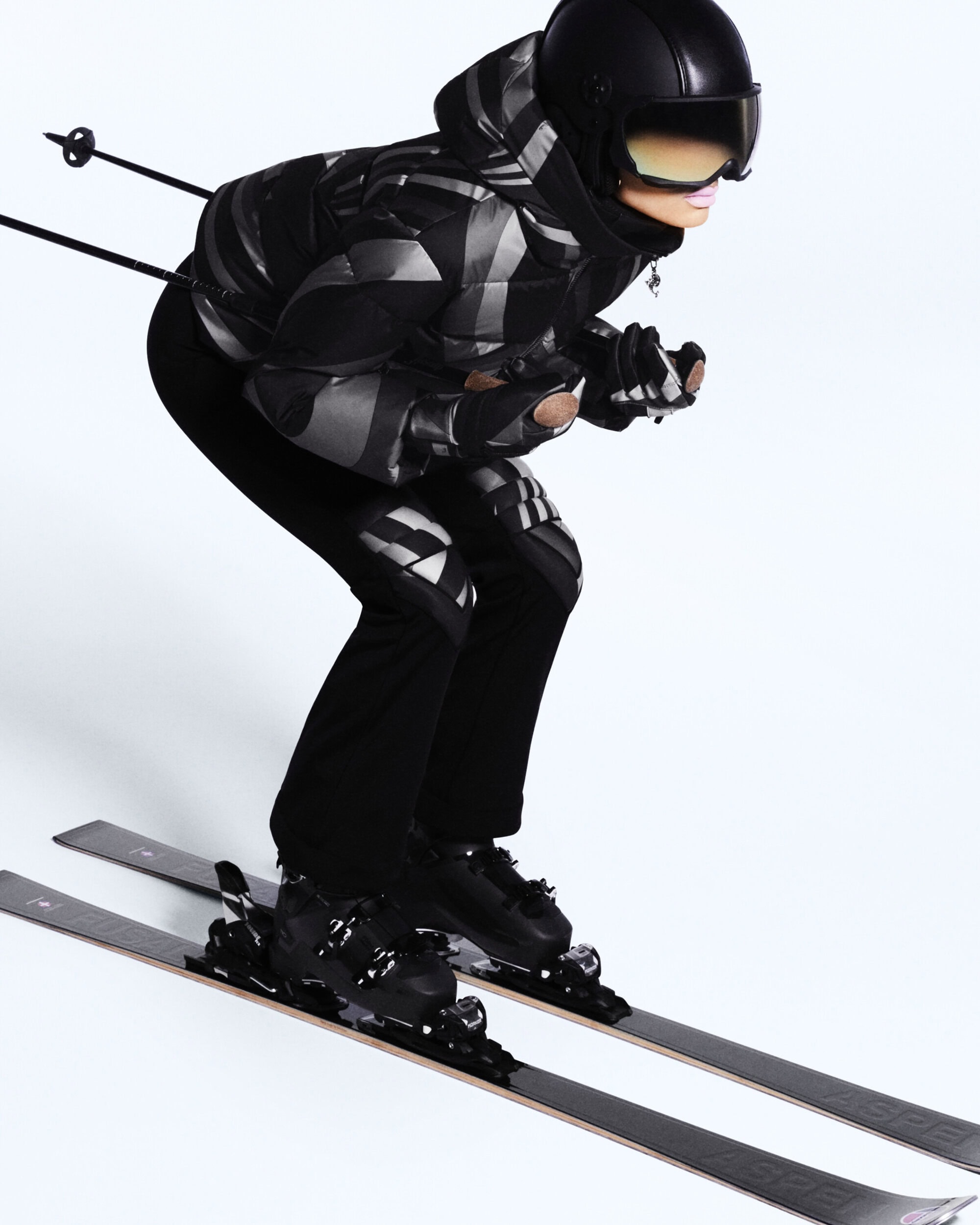 A skier in a black and grey patterned jacket and black ski pants on skis with ski poles against a white background in a studio shot.