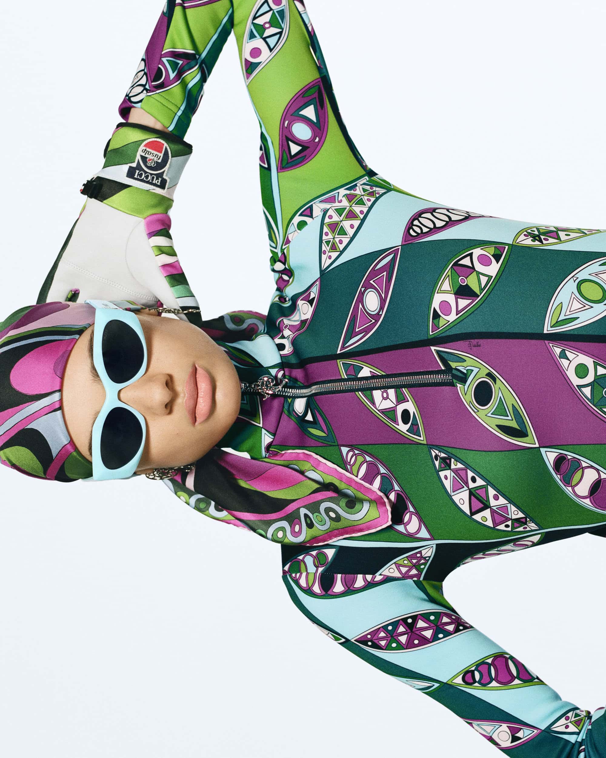 Woman in patterned ski suit and baby blue sunglasses poses against a white background, showcasing a retro fashion style.