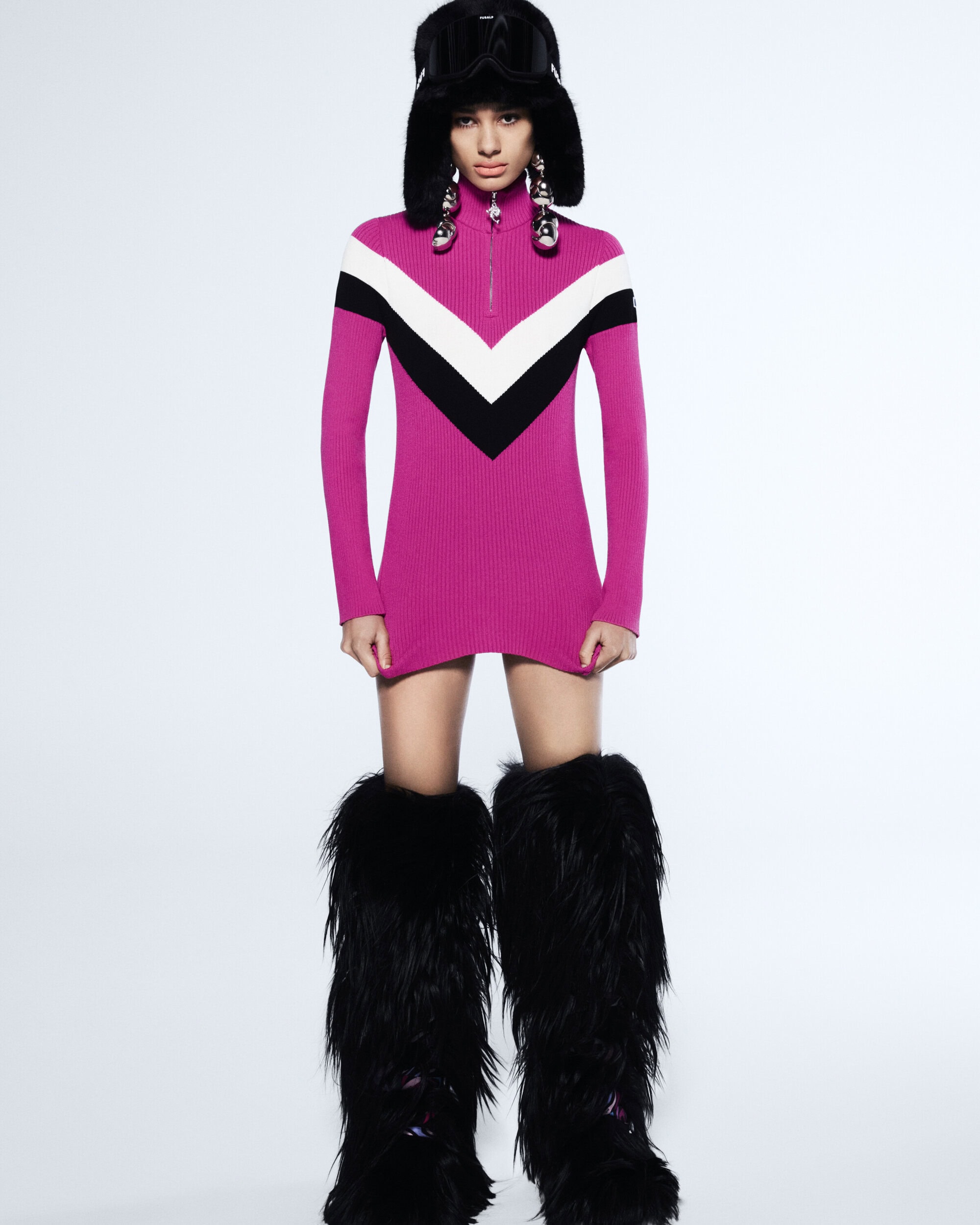 Fashionable woman in a pink mini dress with black and white accents, fur hat with goggles, and furry boots poses in a studio.