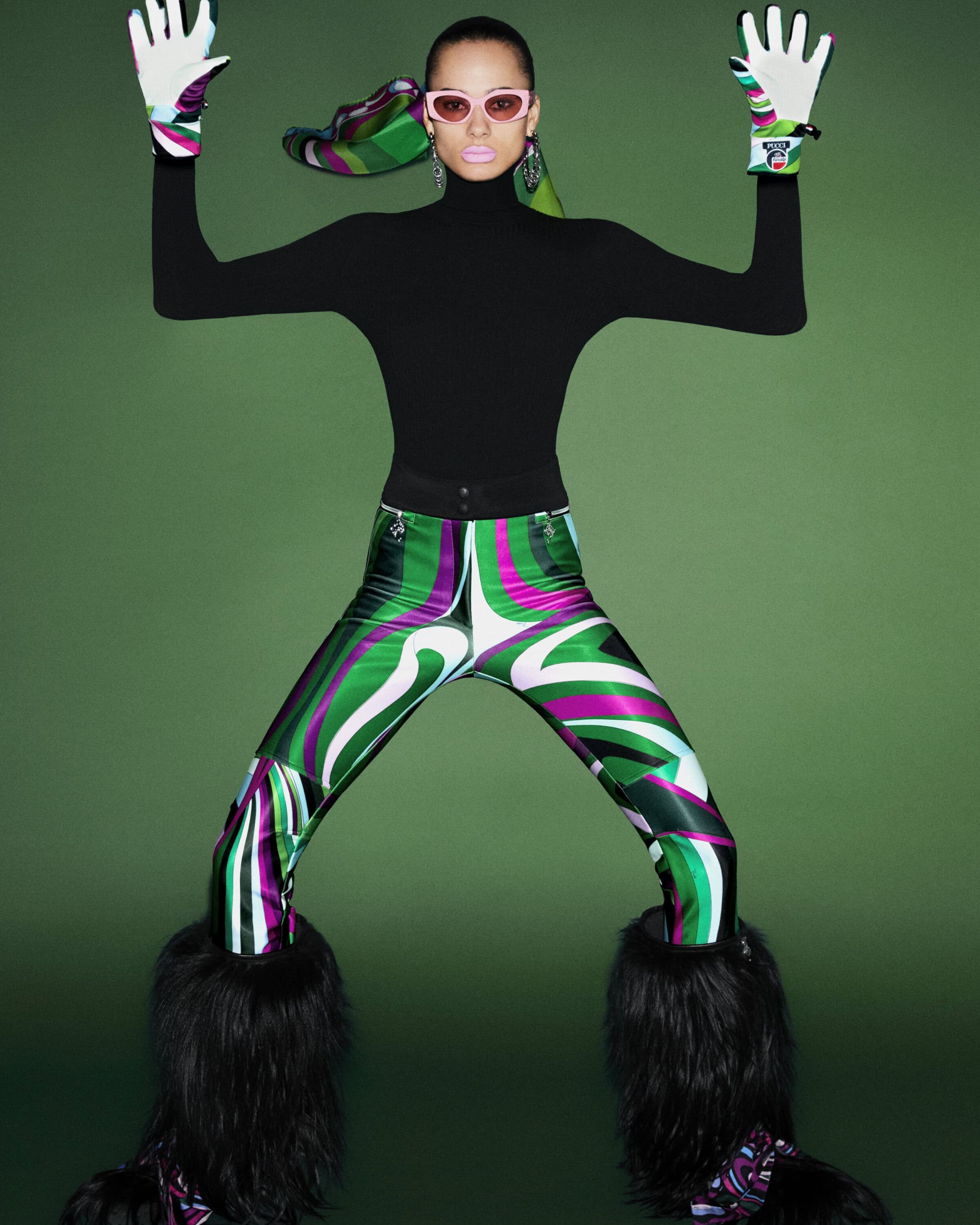 Fashion model in a black turtleneck, patterned pants, furry boots, gloves, and pink glasses posing in a studio setting with a green background.