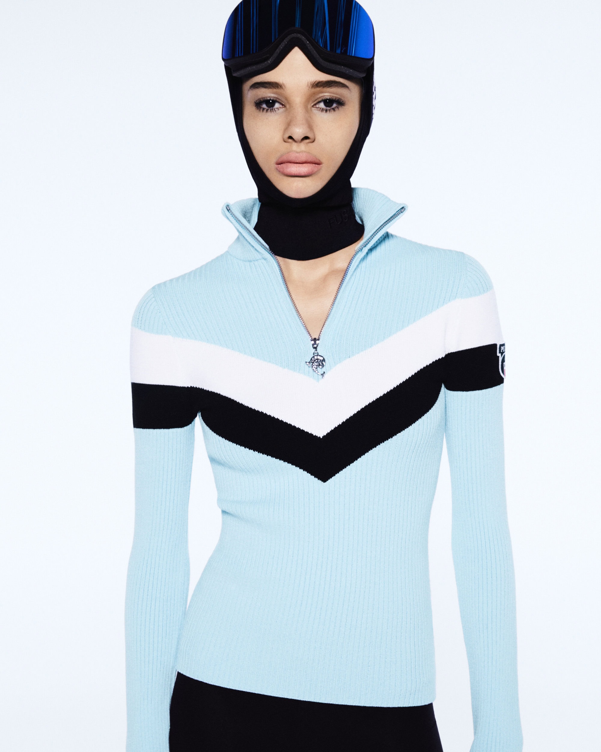 Woman in ski goggles and a black balaclava, wearing a light blue, black, and white chevron-patterned sweater with a partial zipper.