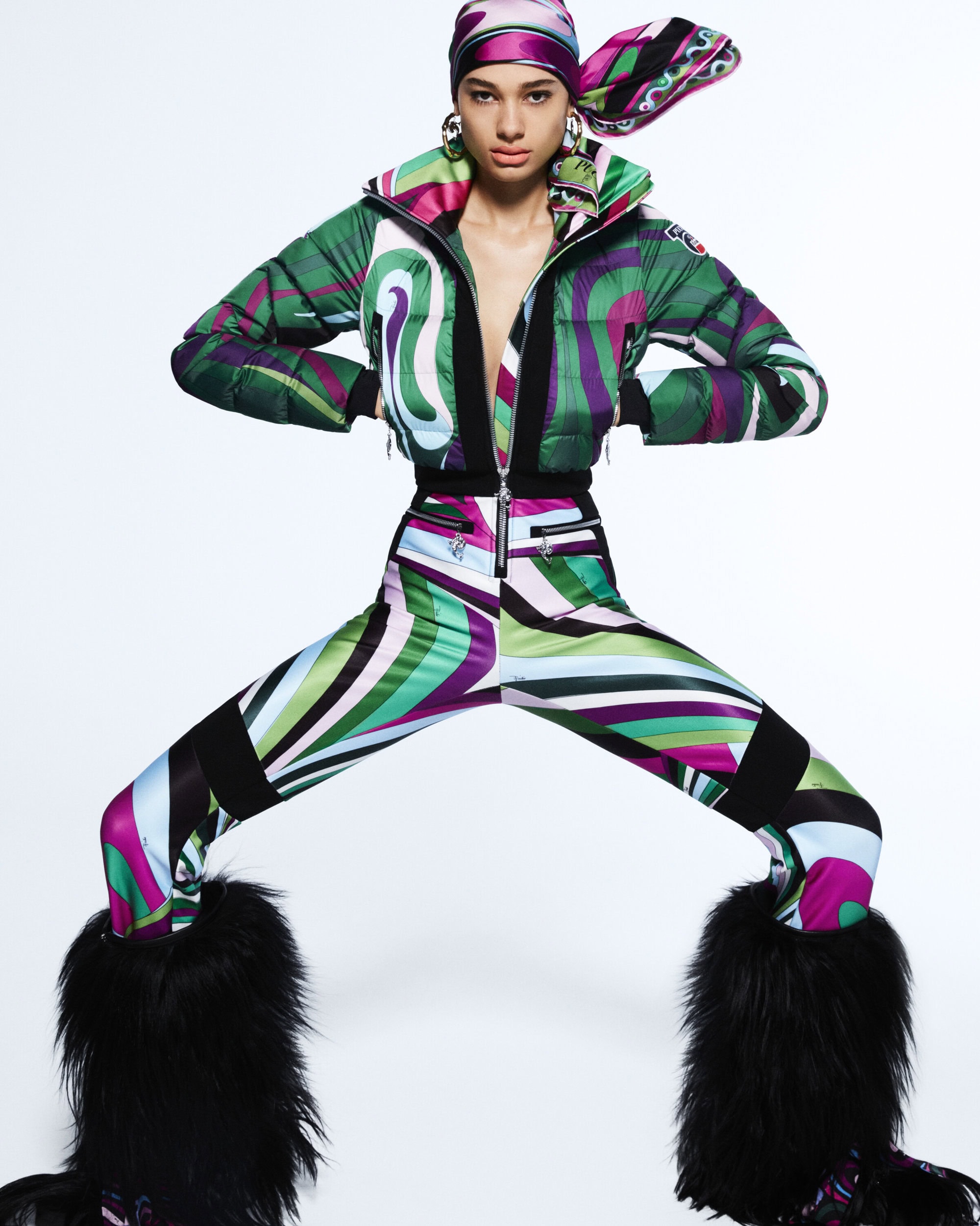 Fashion model in a patterned puffer jacket, pants, headdress, and furry boots poses in a full shot against a white backdrop.