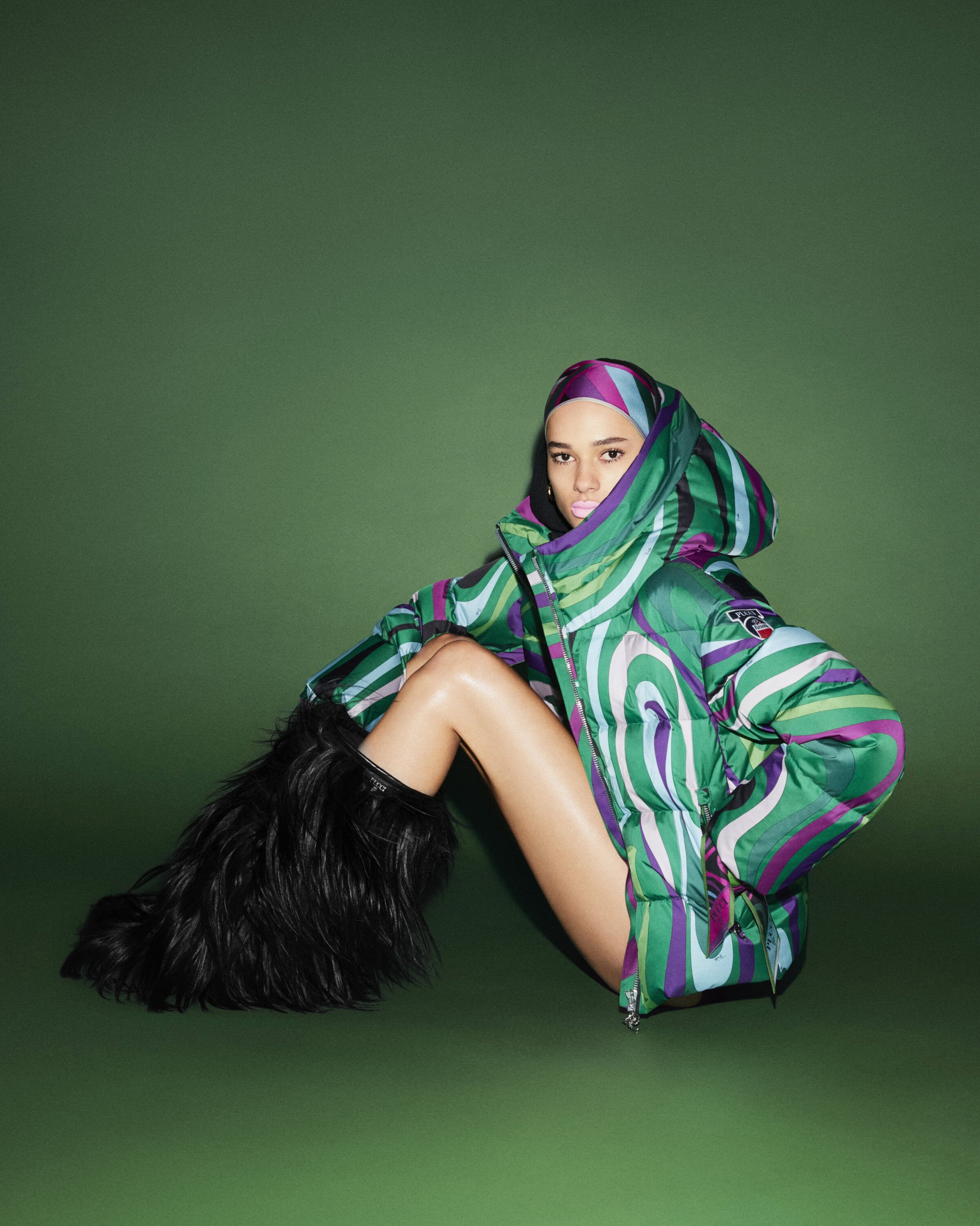 A fashion model in a green patterned puffer jacket poses on a green background, wearing black fur boots and matching headscarf.