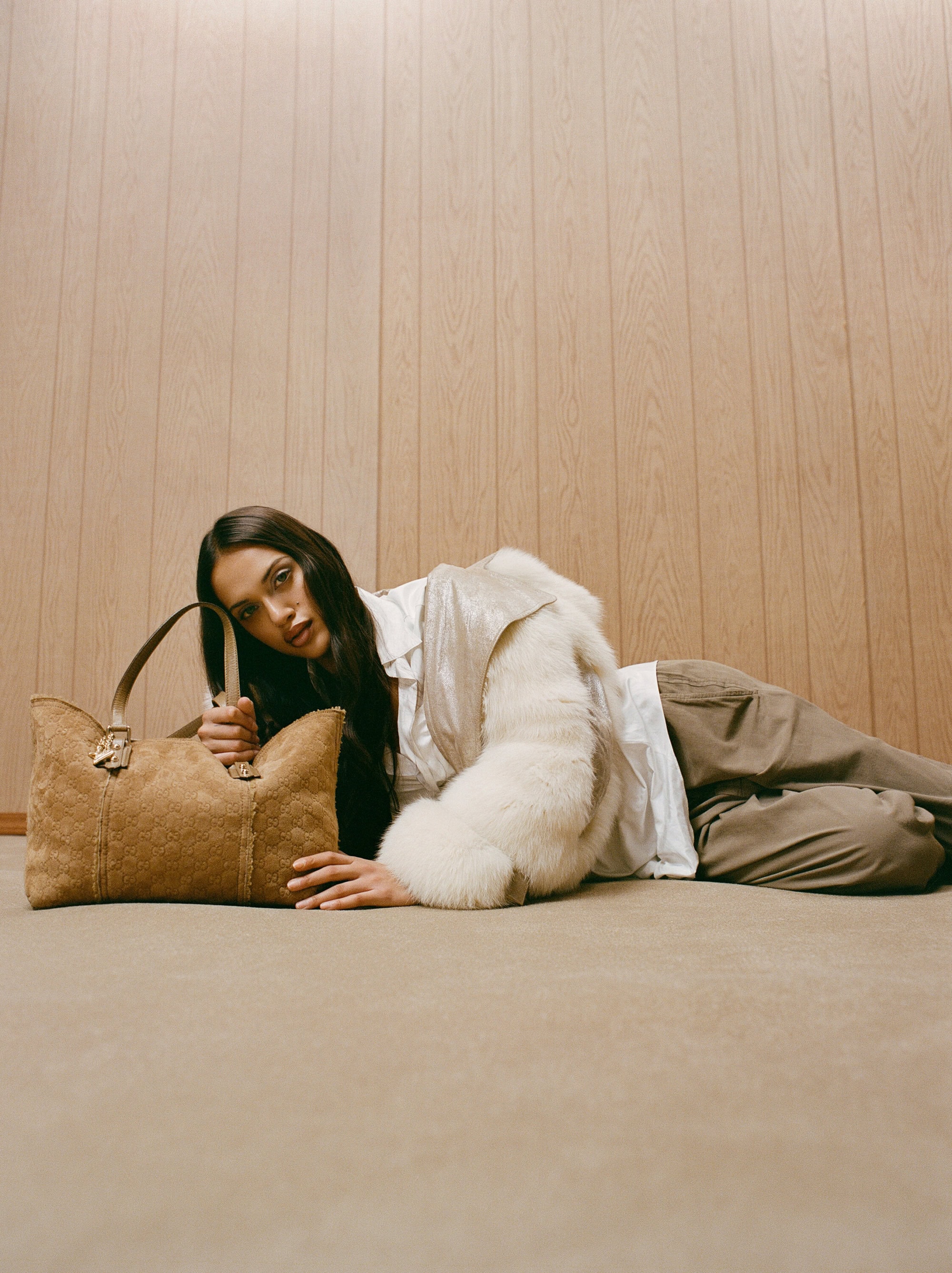 Brunette fashion model with a handbag laying on the floor in front of a wooden wall wearing a jacket with fur sleeves and light-colored pants.