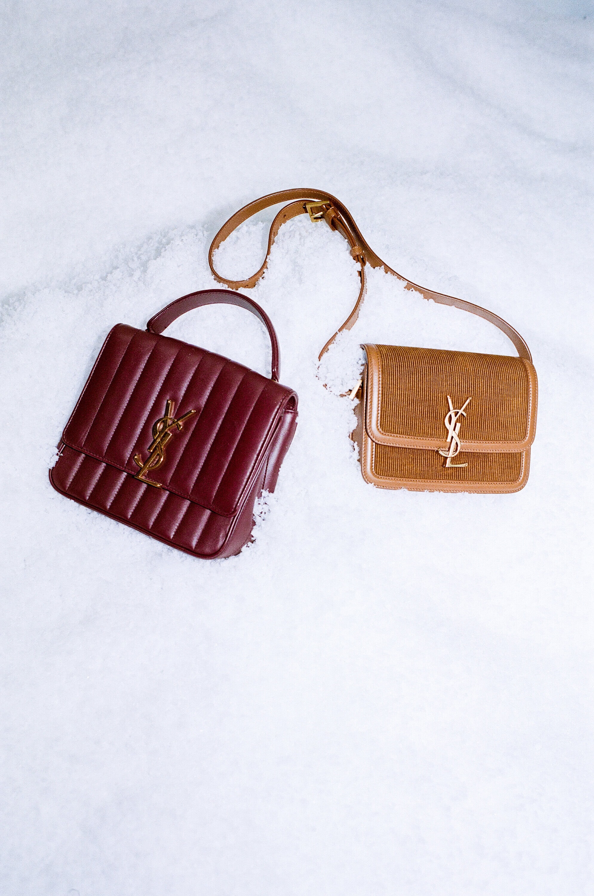 Two Saint Laurent bags, one burgundy with a top handle and one brown with a strap, sit on a surface covered with snow.