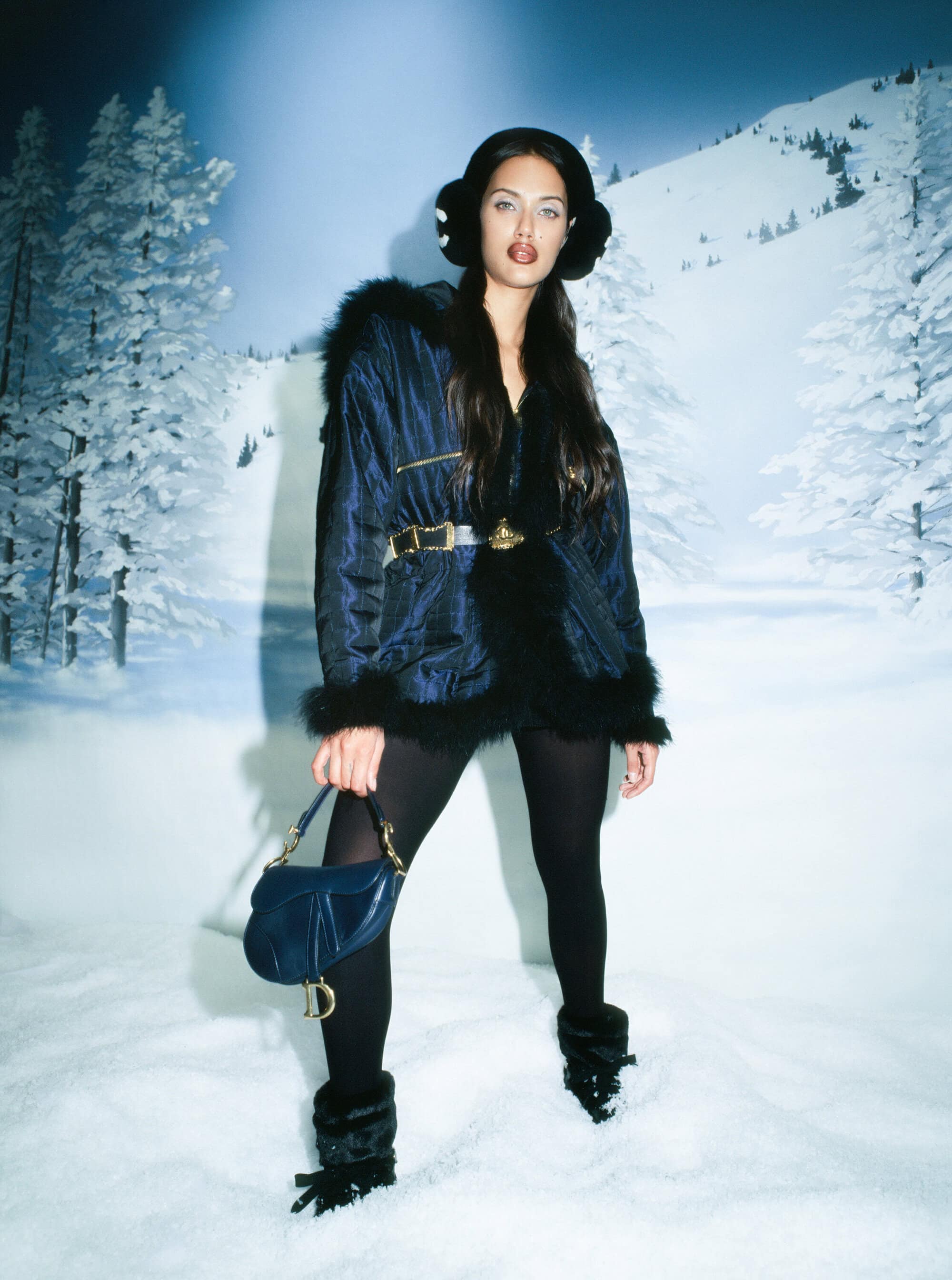 A woman in a fur-lined jacket and earmuffs poses against a snow scene backdrop, holding a blue Dior handbag in a full shot.
