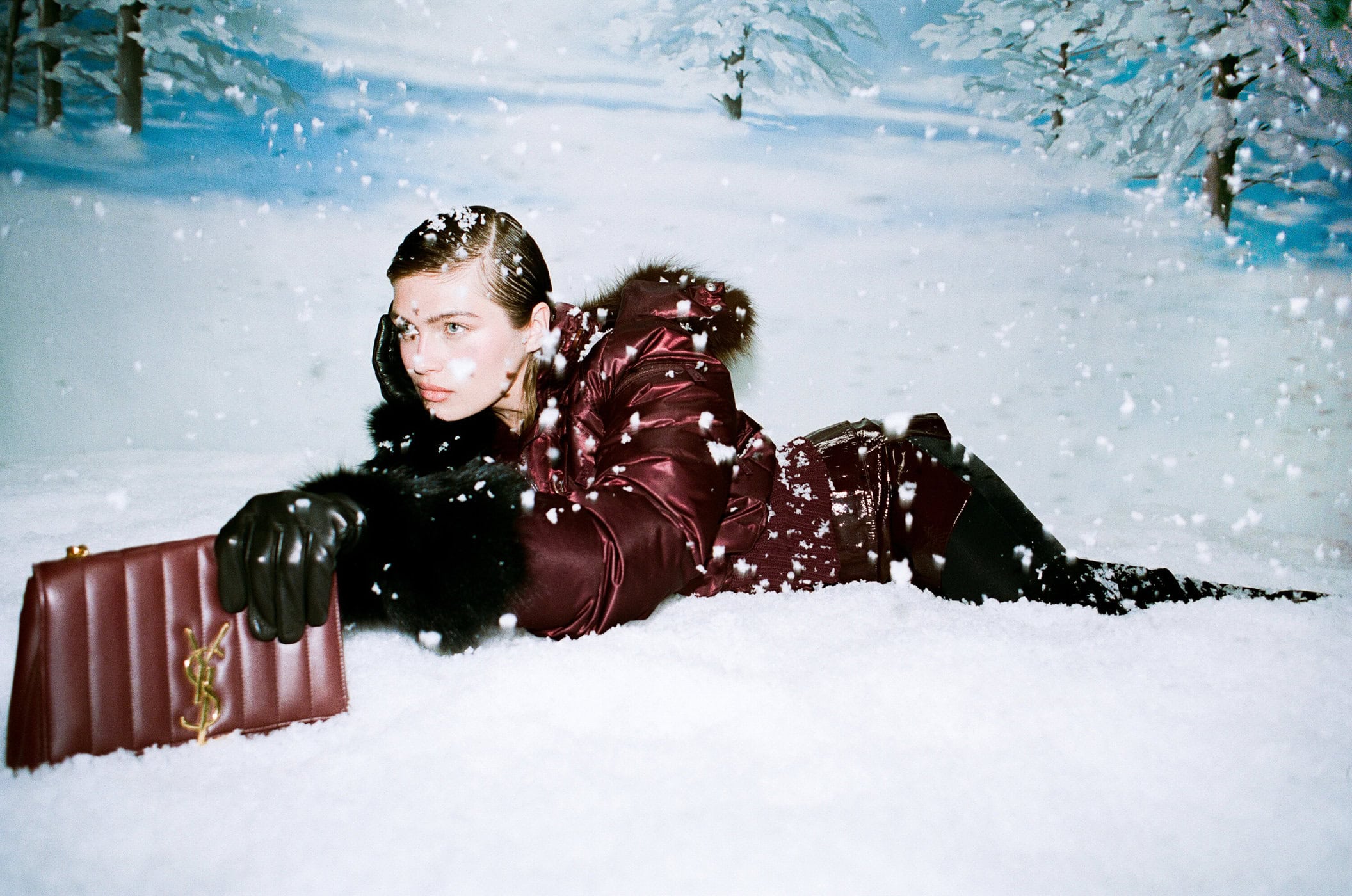 A woman with a burgundy coat and black gloves lays in the snow holding a designer purse; trees and snow in background.