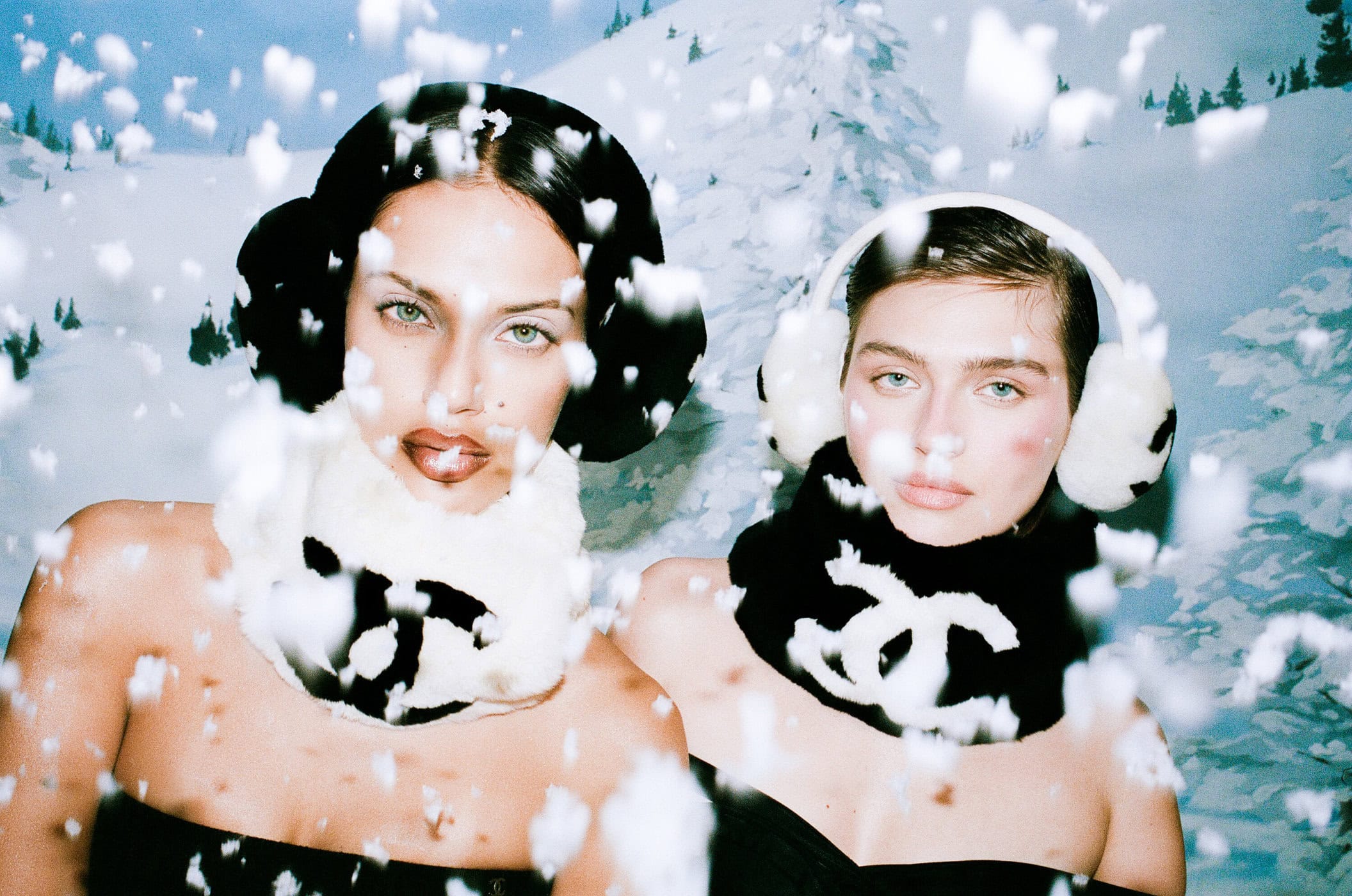 Two women in winter fashion against a snowy mountain backdrop, adorned with Chanel accessories and falling snowflakes, showcase cold weather style.