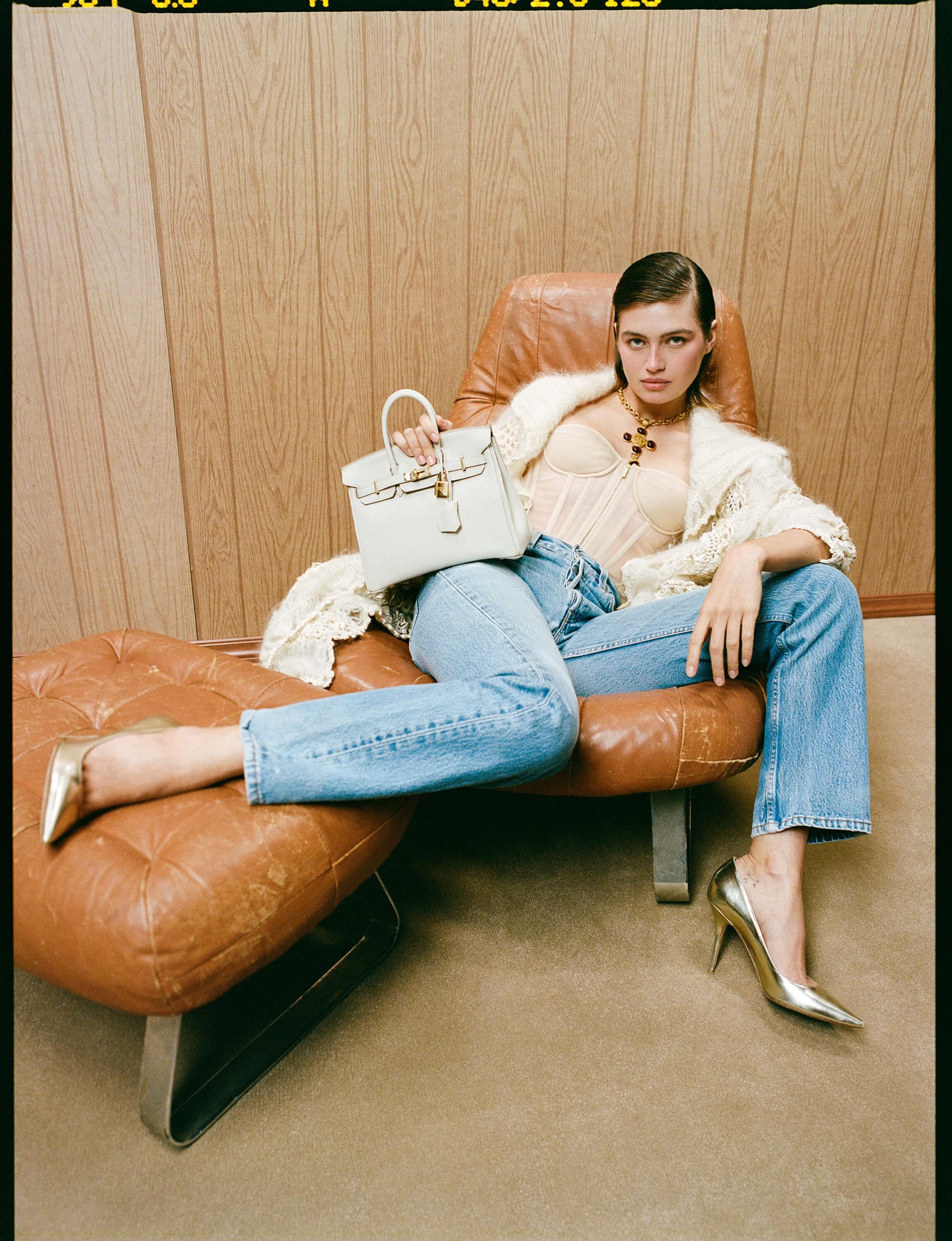 Model Dalianah Arekion posing in an armchair, holding a light handbag and wearing blue jeans and metallic heels.