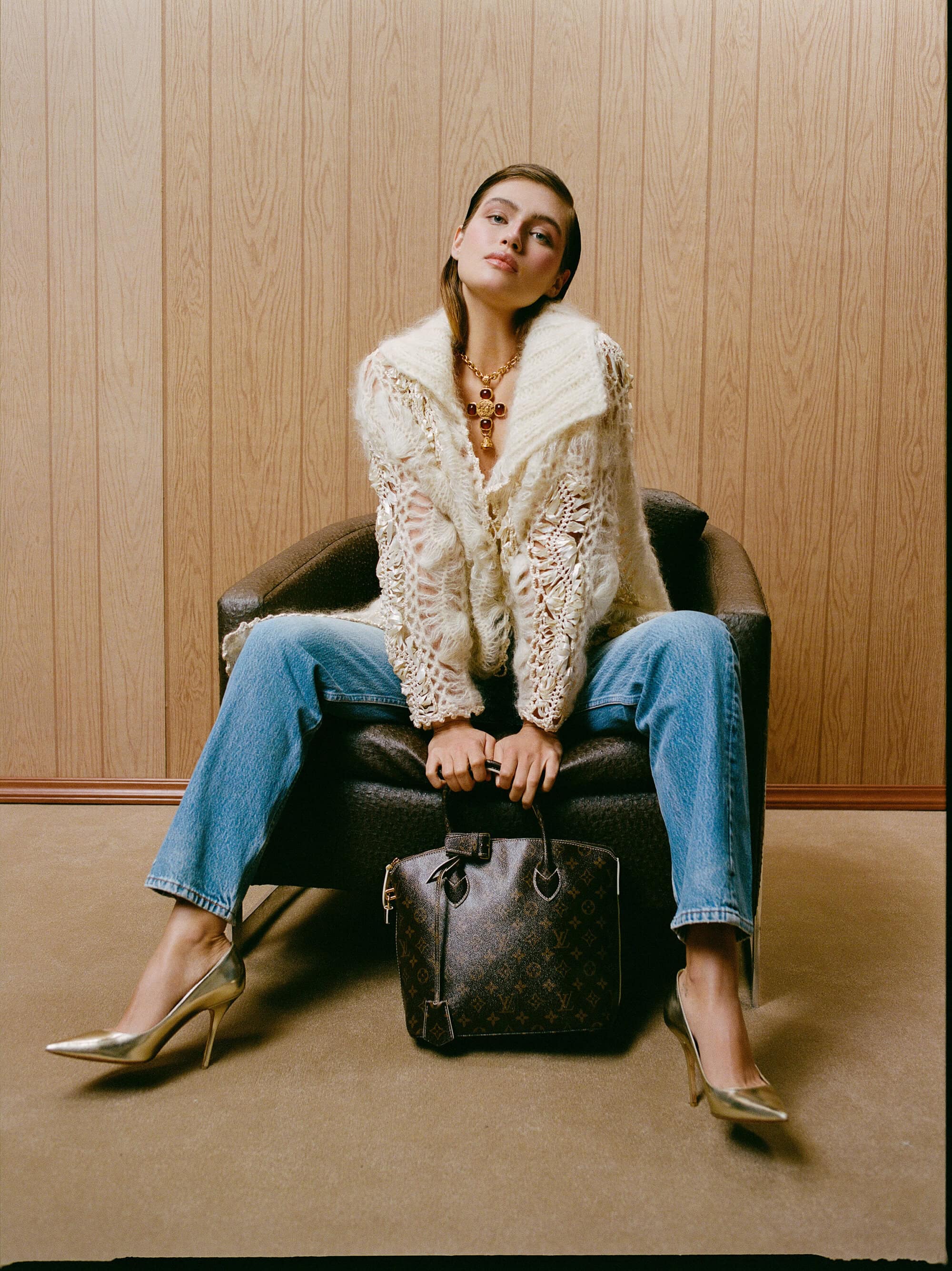 Model Dalianah Arekion seated in an armchair wearing a cream knit cardigan, blue jeans, and gold heels with a monogram handbag.