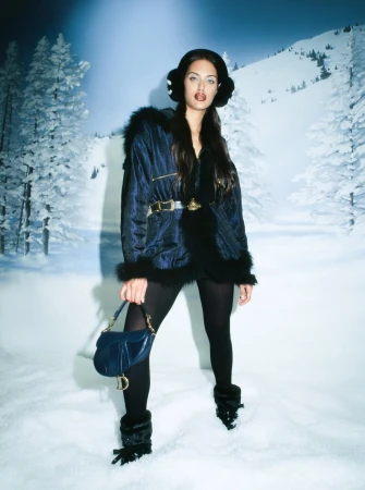 Model wearing dark blue winter coat and holding a blue handbag against a snowy winter backdrop