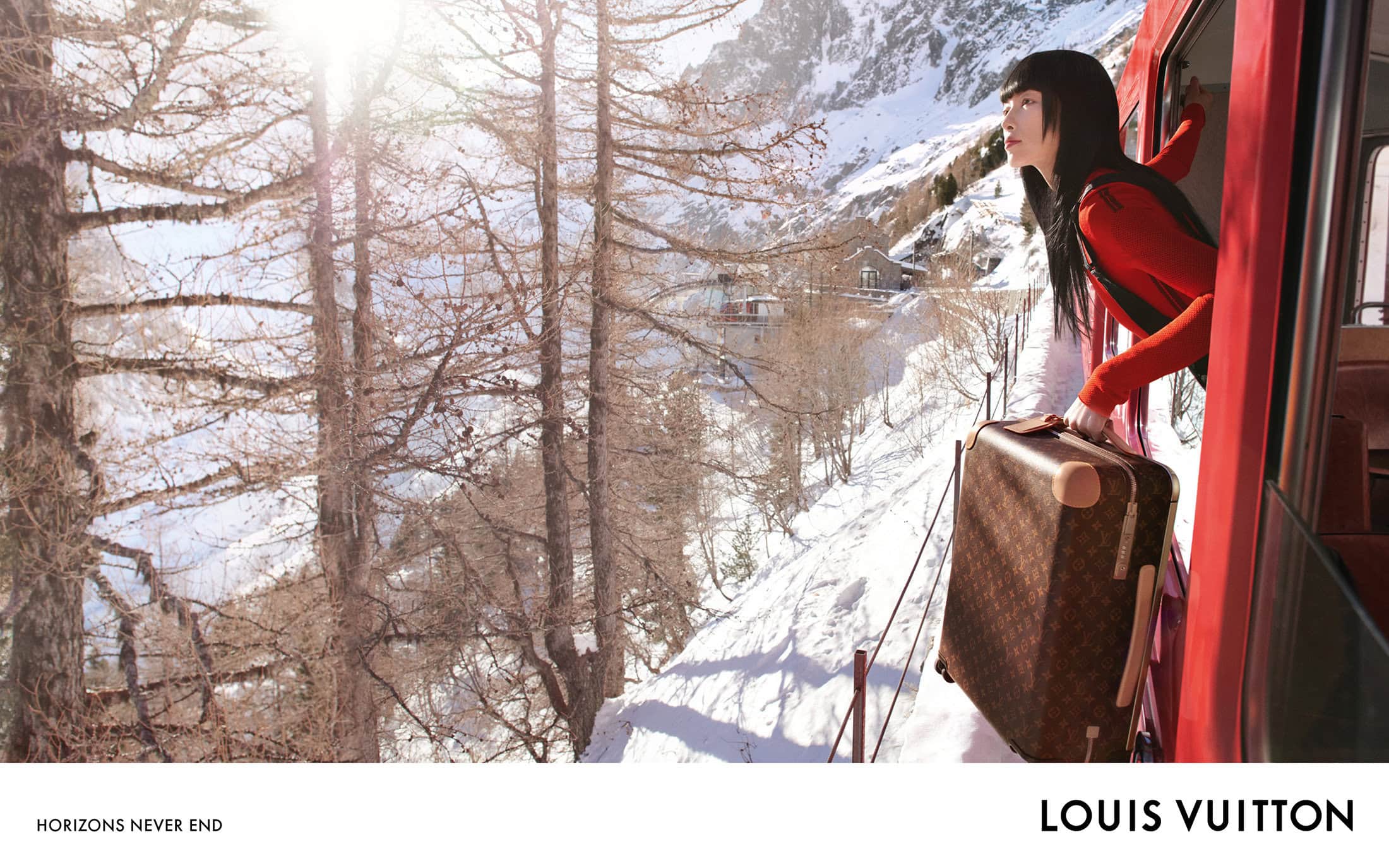 Woman gazes from a red train in snowy mountains holding a Louis Vuitton suitcase; evergreen trees and bright sunlight enhance this travel scene.