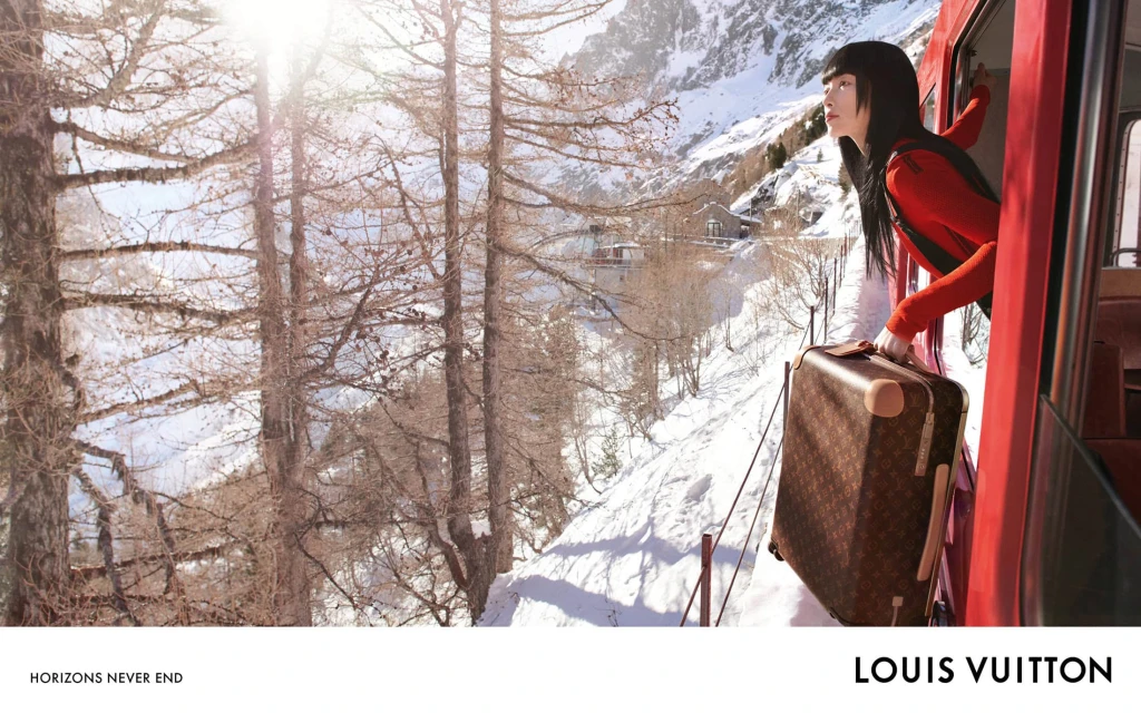 Woman gazes from a red train in snowy mountains holding a Louis Vuitton suitcase; evergreen trees and bright sunlight enhance this travel scene.