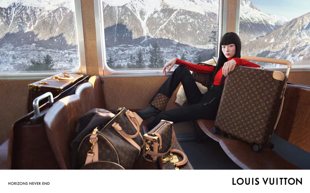 A woman poses with Louis Vuitton luggage inside a train car with a snowy mountain range visible through the window in the background.