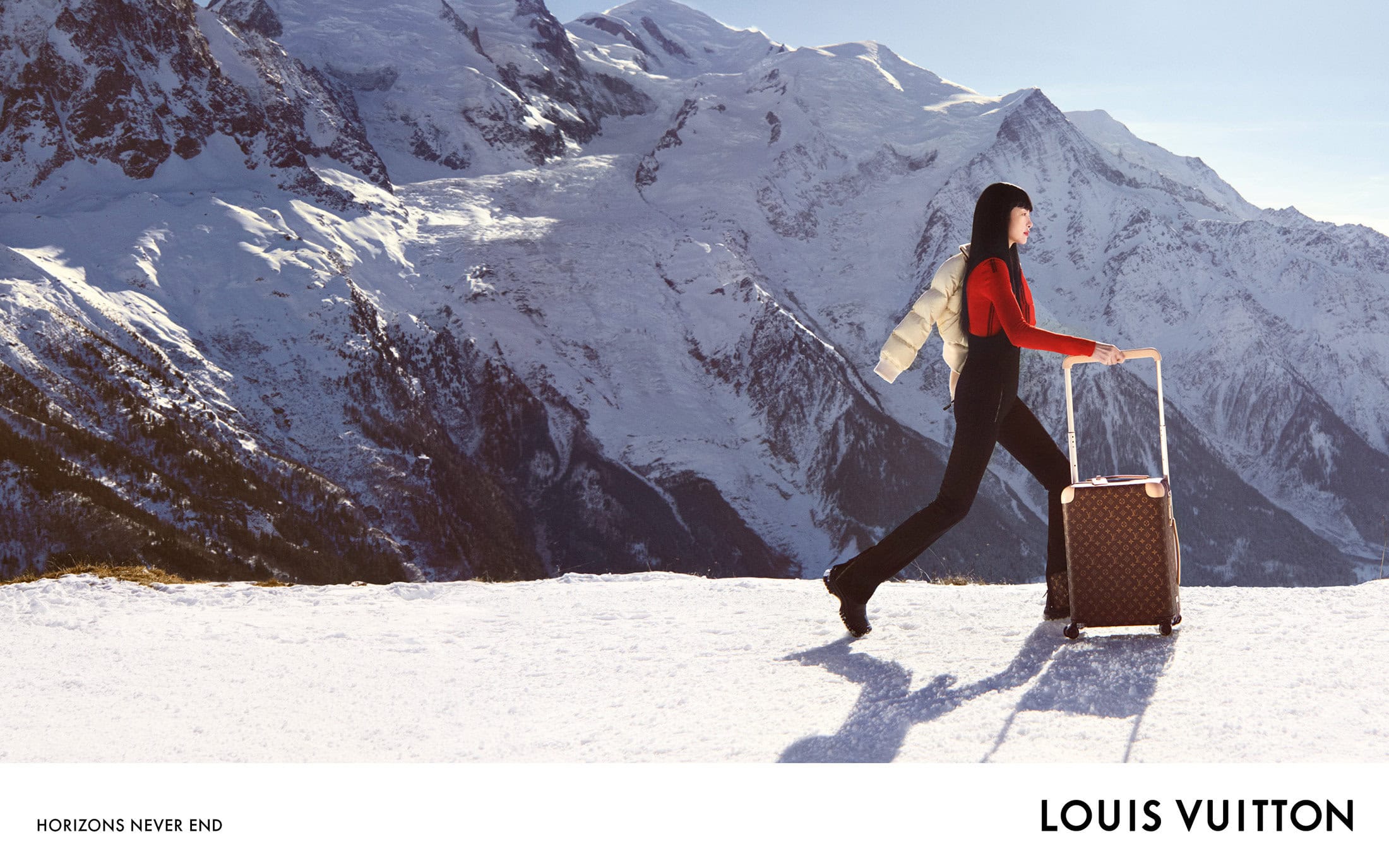 A woman in a red sweater and black pants walks in a snowy landscape pulling a suitcase, with a snowy mountain range in the background.