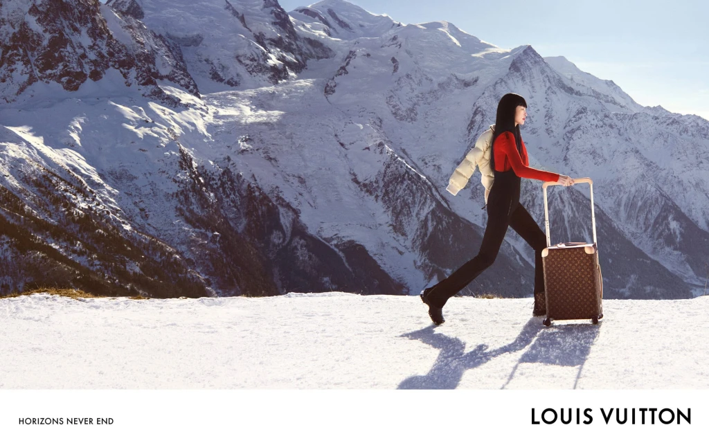 A woman in a red sweater and black pants walks in a snowy landscape pulling a suitcase, with a snowy mountain range in the background.